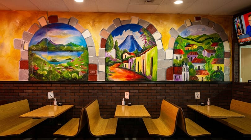 Restaurant interior with wooden booths, brick wall, and a colorful mural depicting three arched landscape scenes.