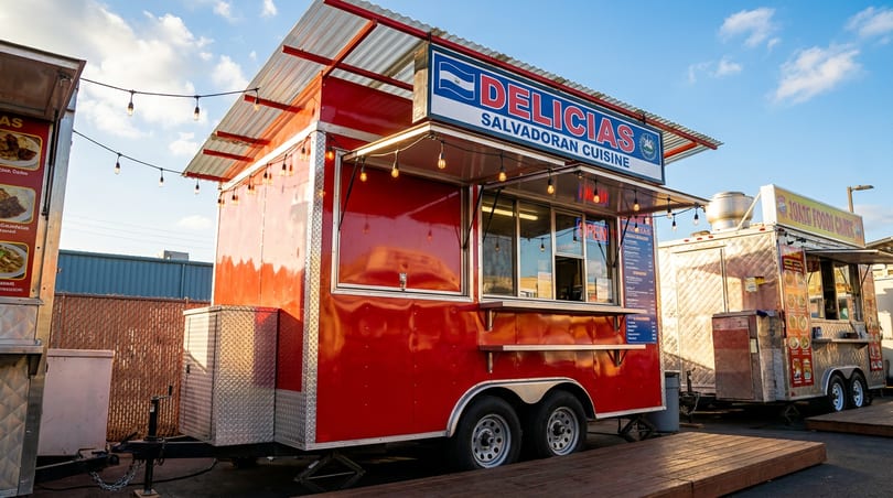 Red "DELICIAS SALVADORAN CUISINE" food truck with string lights, next to other food trucks outdoors.