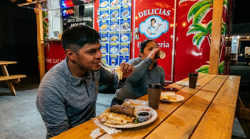 Two diners enjoying cheesy food and drinks at a vibrant red food truck at night.