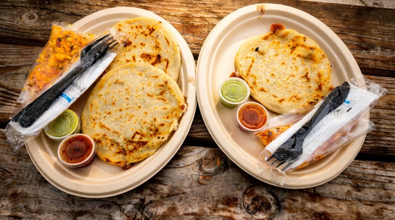 Two plates of golden pupusas with green and red sauces, plus cutlery, on a wooden surface.