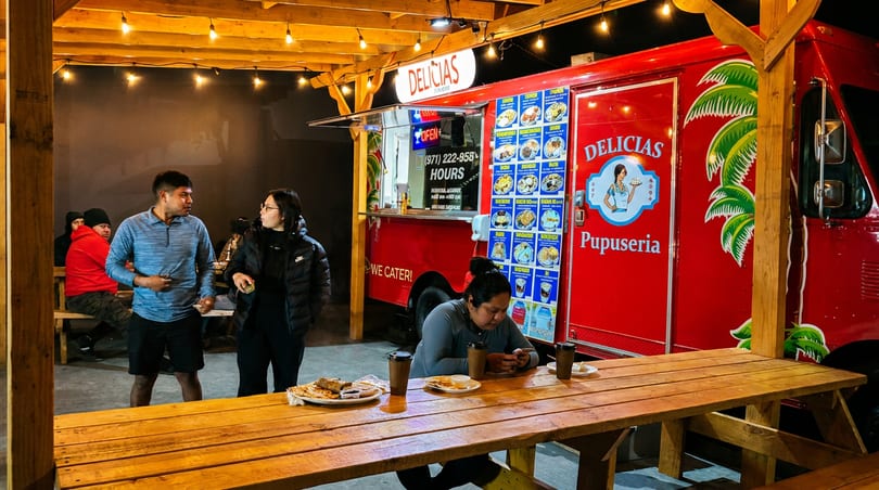 People gather at night around a red Delicias Pupuseria food truck with string lights and picnic tables.